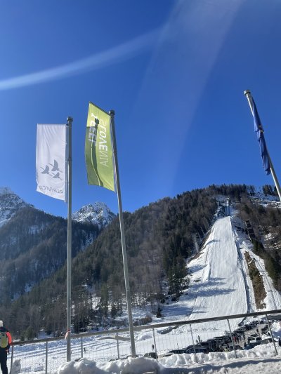 Planica zame ni samo dolina med gorami – je občutek.Vsakič, ko pomislim na Planica, mi v ušesih še vedno zazveni navijanje. Mrzel zrak, para iz ust, zastave, ki plapolajo, in tisti poseben trenutek tišine tik preden skakalec poleti. Potem pa eksplozija veselja, kriki, aplavz – kot da diha cela dolina z nami.Spomin na tekme je prepleten z glasovi ljudi, s smehom, s toplimi čaji v rokah in z občutkom pripadnosti. Tam nisem bil/a samo gledalec – bil/a sem del množice, del zgodbe, del navijaškega srca, ki bije za pogumne polete.Poletna Planica ima čisto drugo energijo. Sonce riše sence po pobočjih, dolina je mirnejša, a nič manj mogočna. Plezanje na velikanko ni samo vzpon po stopnicah – je soočenje s samim seboj. Vsak korak je malo težji, noge pečejo, srce razbija, a na vrhu… na vrhu je razgled, ki ti vzame besede. Takrat razumeš, kako noro pogumni so tisti, ki od tam skočijo v praznino.In potem je še Tamar. Pot do Tamarja je sprehod v tišino. Gozd šumi, pot škripa pod nogami, v glavi se naredi prostor. Tam čas teče počasneje. Tam se spomini ne derejo – samo mirno obstajajo.Planica, tekme, navijanje, poletna dolina, vzpon na velikanko in Tamar – vse to je del mene. Del mojih korakov, mojega glasu v množici in mojega miru v gorah.To niso samo kraji. To so občutki, ki jih nosim s sabo. 